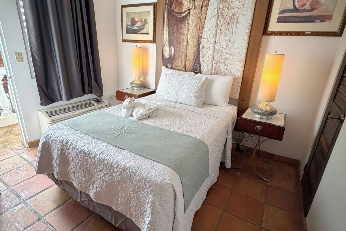 A neatly made bed with a white and green bedspread, two bedside lamps, and a large painting above the headboard.