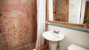 Une salle de bain avec un mur de douche en pierre, un lavabo blanc, un miroir et des toilettes.