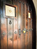 Una puerta de madera con diseño antiguo, que incluye una placa en relieve, elegantes pomos de metal y un pequeño ventanal.
