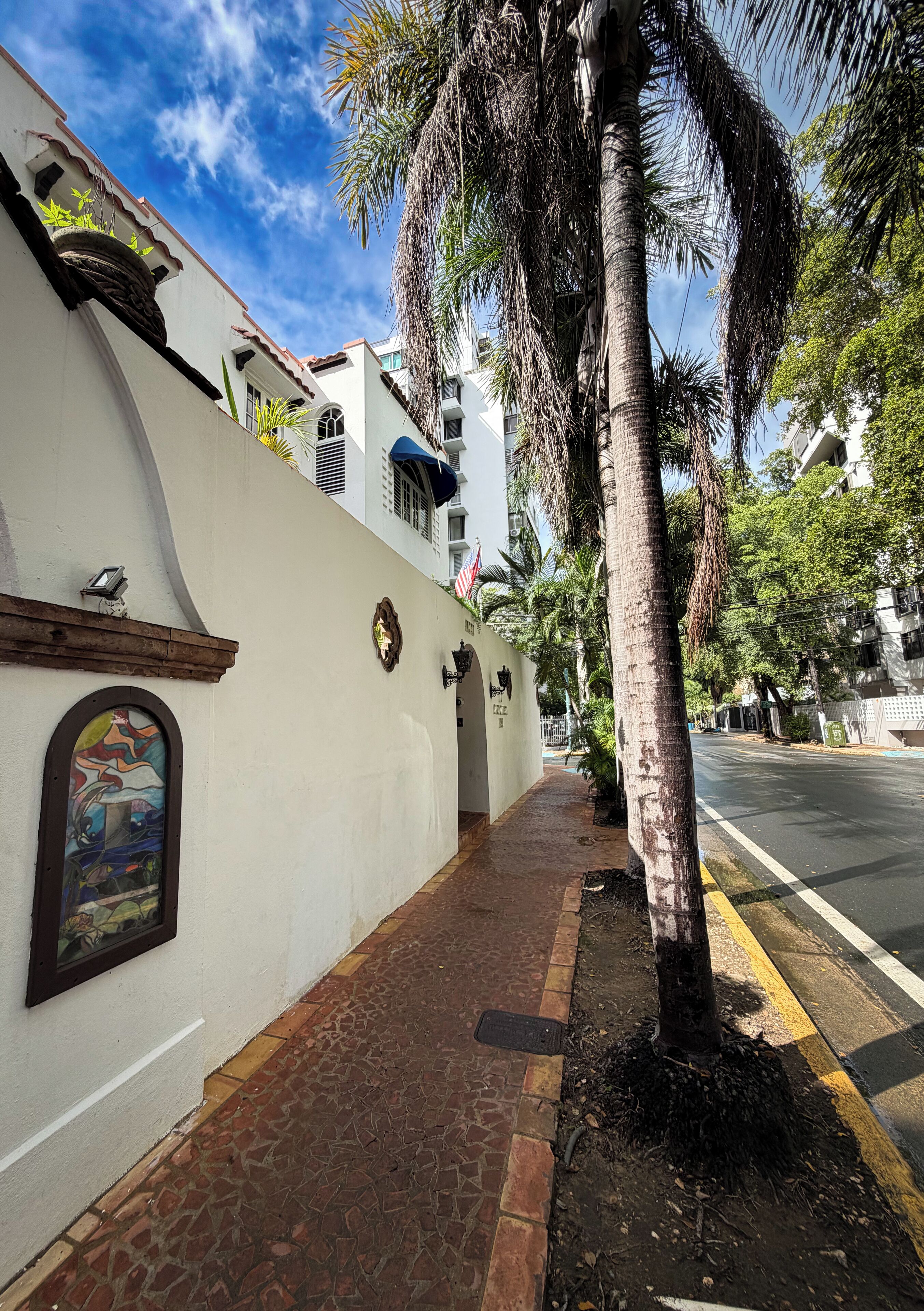 A sidewalk with a palm tree, a white building with a mural, and a clear blue sky.
