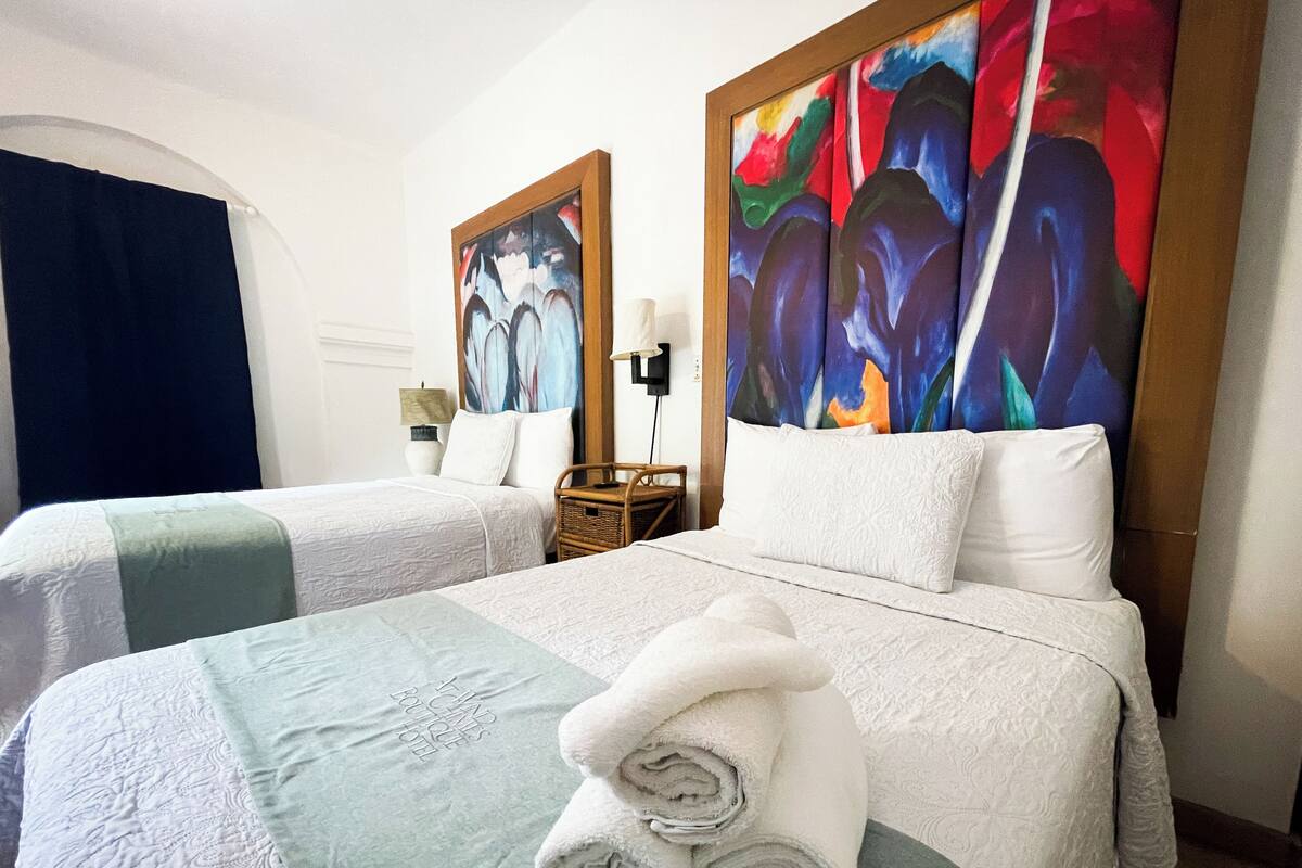 A hotel room with two beds, a large abstract painting above each bed, a folded towel on the bed, and a lamp on a nightstand.