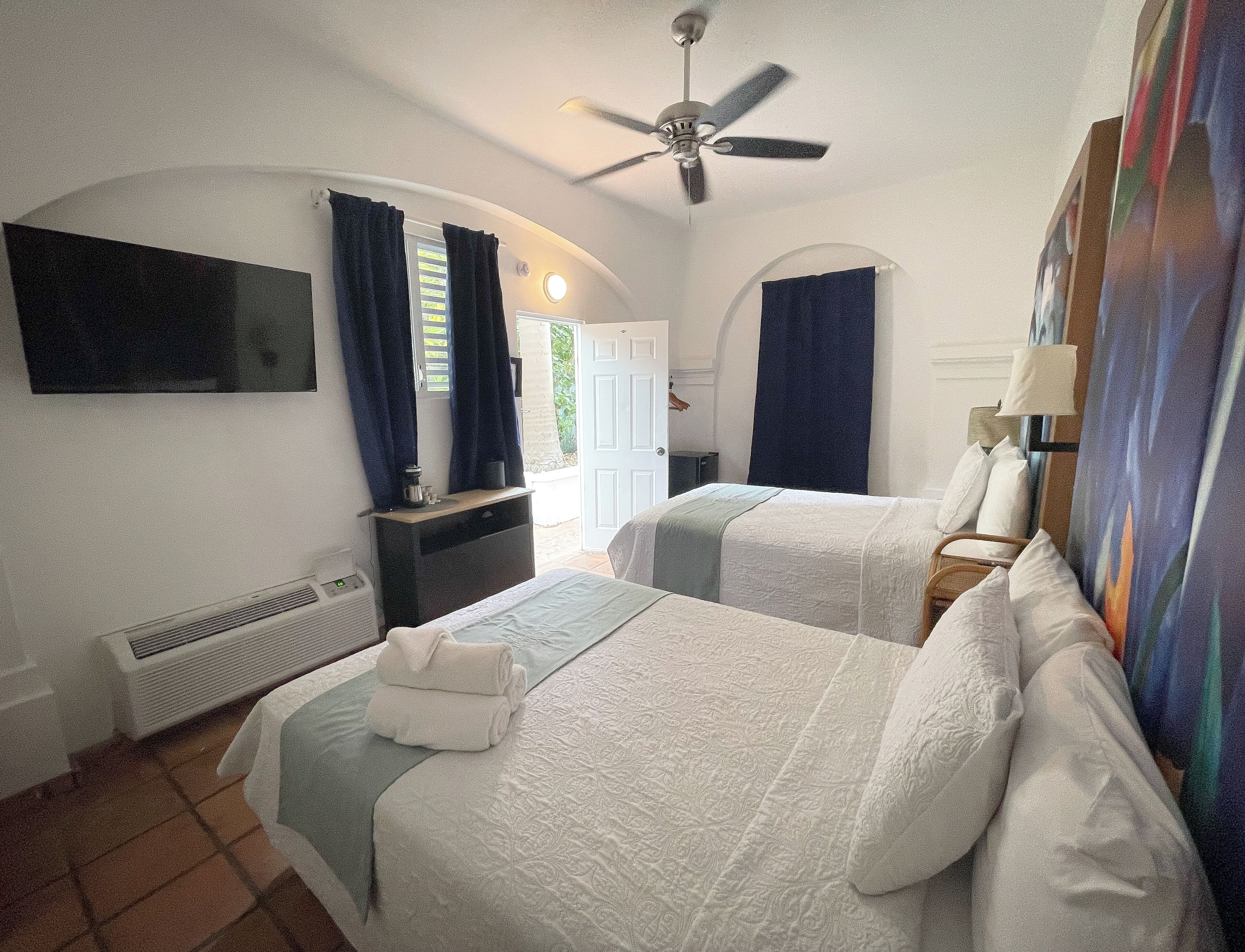 Ein Hotelzimmer mit zwei Betten, einem Deckenventilator, einem an der Wand montierten Fernseher und einem Fenster mit Vorhängen.