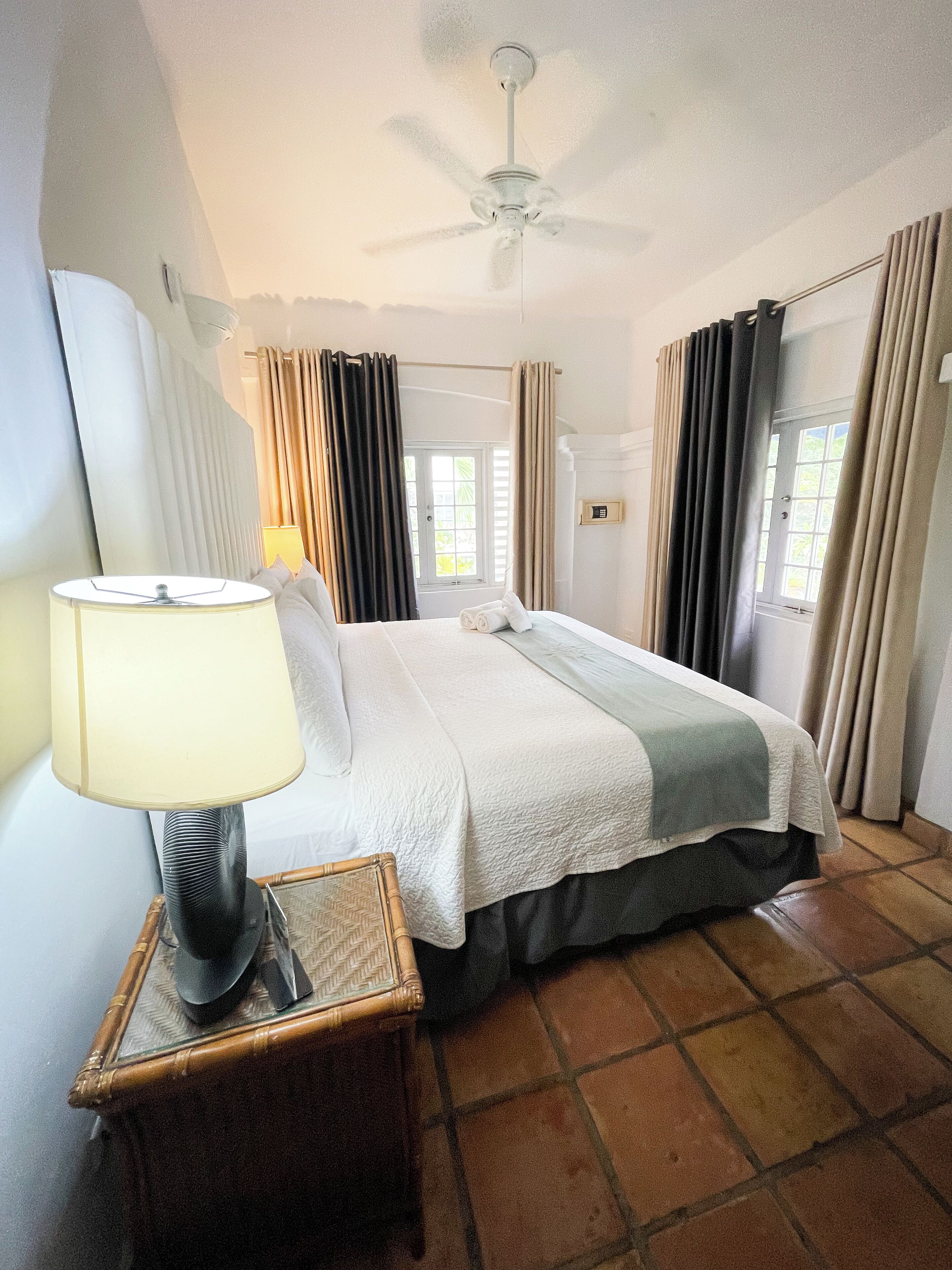 A hotel room with a bed, a bedside table with a lamp, a ceiling fan, and a window with curtains.