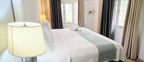 A hotel room with a bed, a bedside table with a lamp, a ceiling fan, and a window with curtains.