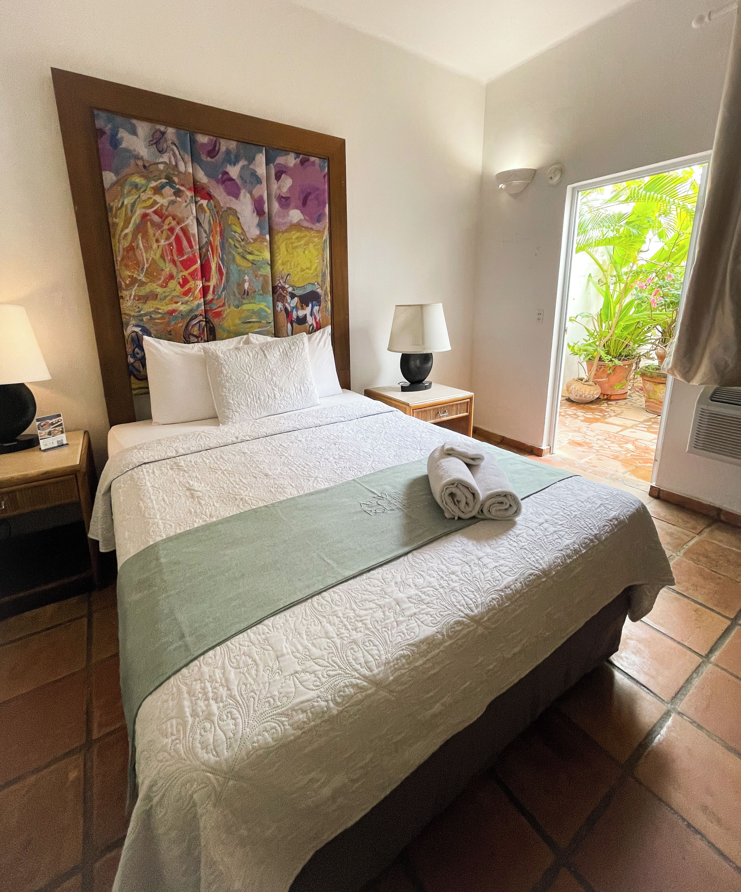 A neatly made bed with a green throw, a painting above it, and a nightstand with a lamp.