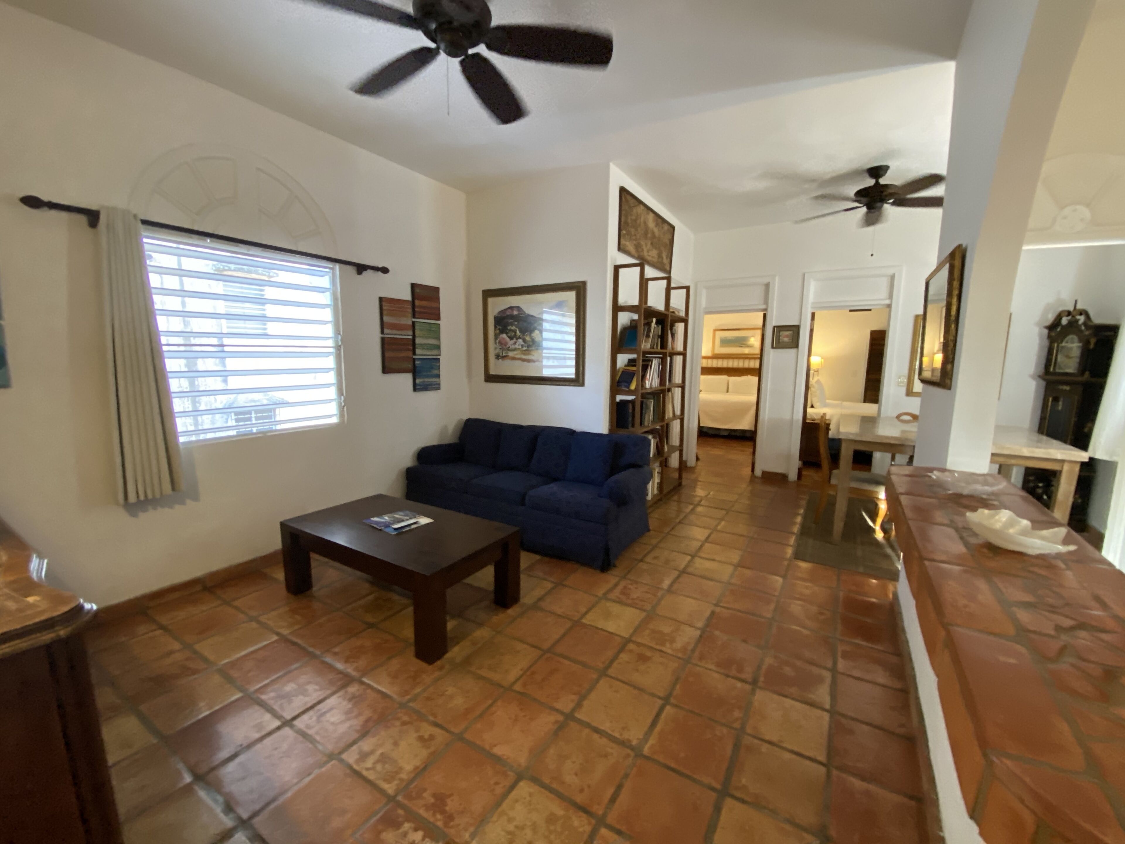 A living room with a blue sofa, a coffee table, and a bookshelf. There are ceiling fans, a window with blinds, and a tiled floor.