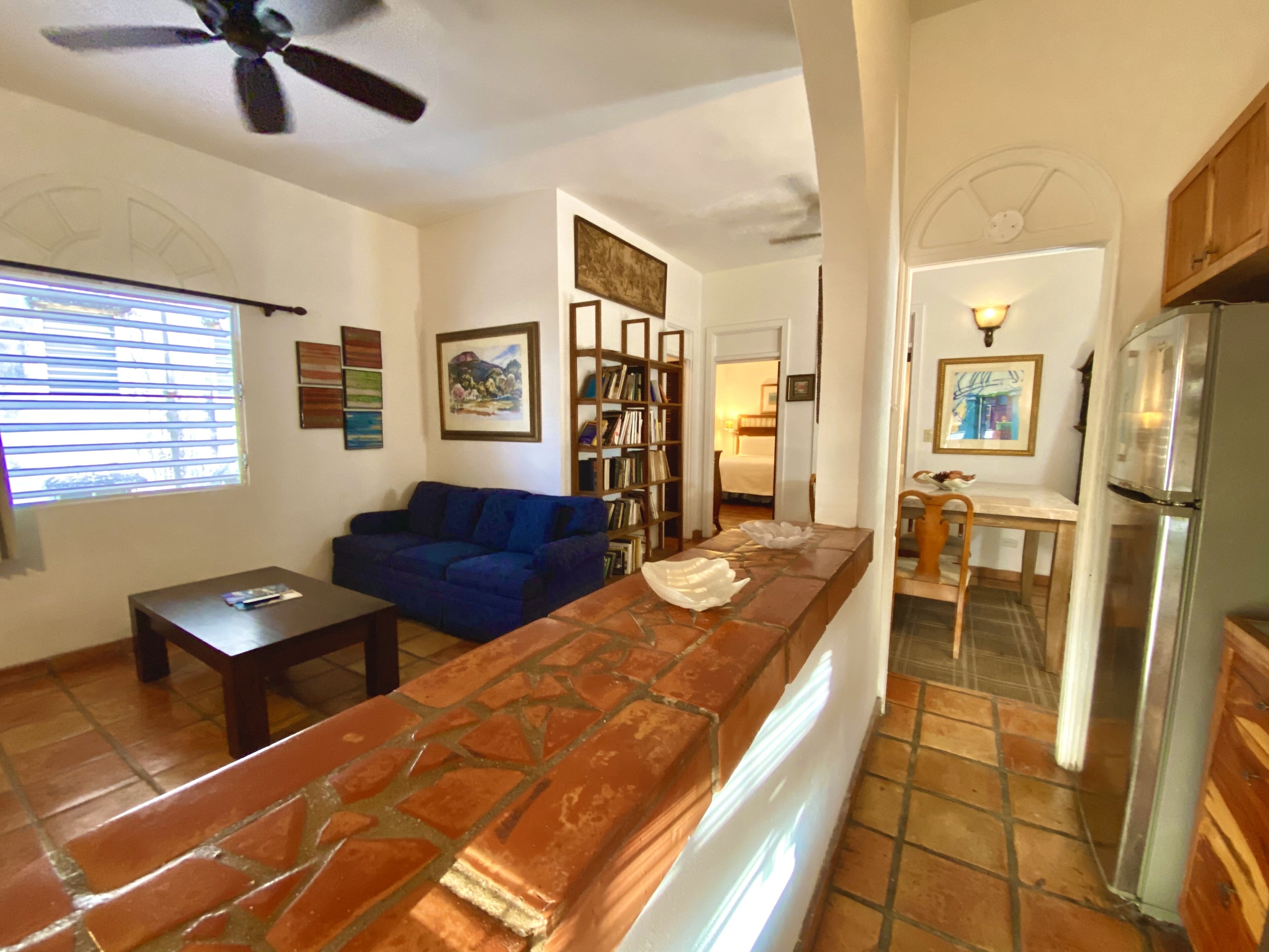 A living area with a sofa, a coffee table, a bookshelf, and a kitchen area with a refrigerator.