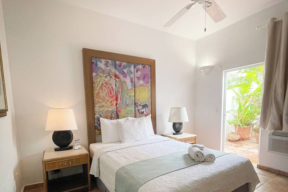 A hotel room with a bed, two bedside tables, a ceiling fan, a window with curtains, and a view of greenery outside.