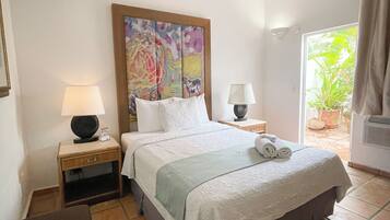 A hotel room with a bed, two bedside tables, a ceiling fan, a window with curtains, and a view of greenery outside.