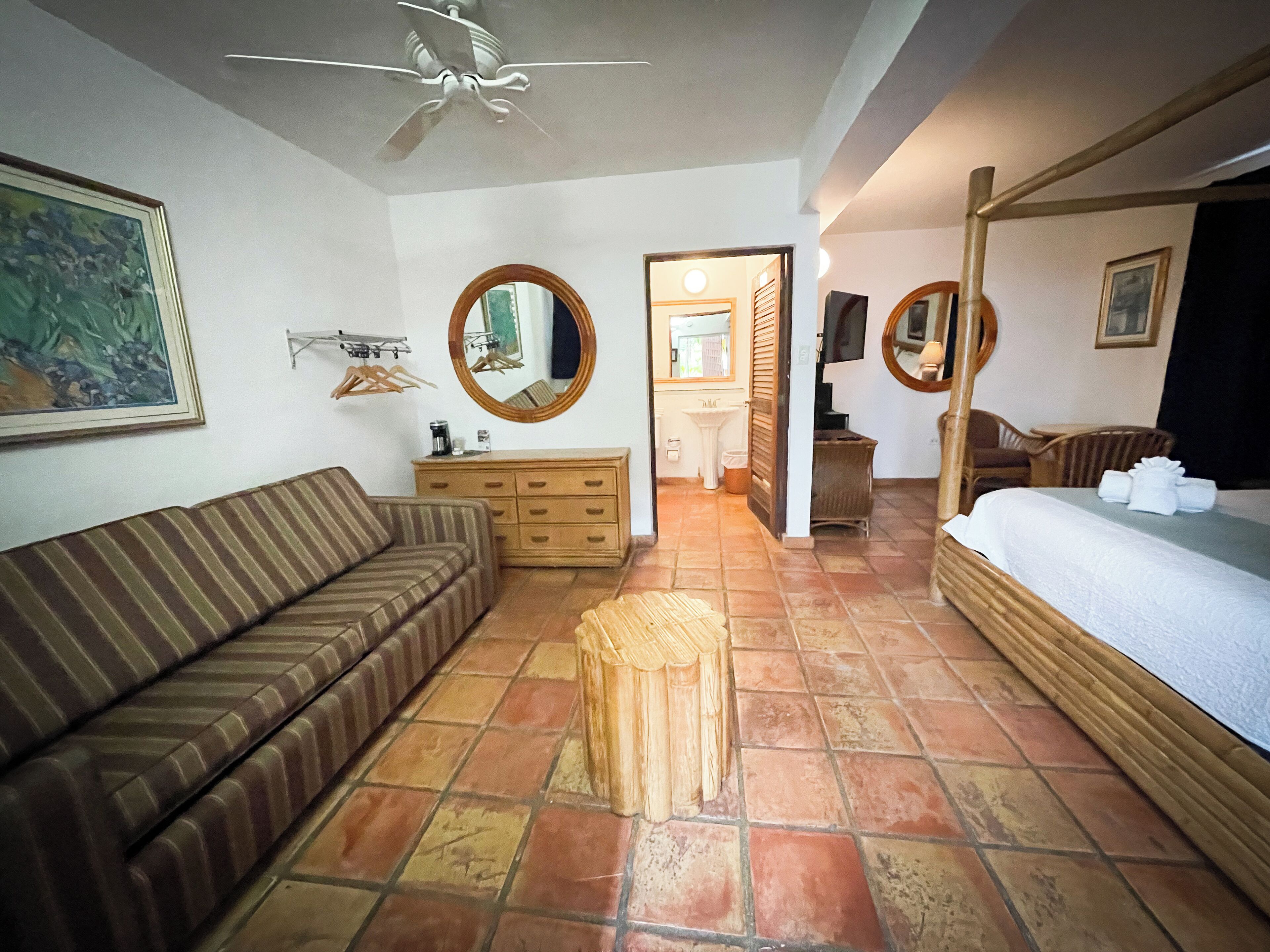 A hotel room with a striped sofa, a wooden dresser, a bed with a white mattress, a round mirror, and a ceiling fan.