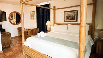 Una cama con dosel, ropa de cama blanca y almohadas, un espejo redondo, una silla de madera, una mesita con una lámpara y un cuadro en la pared.
