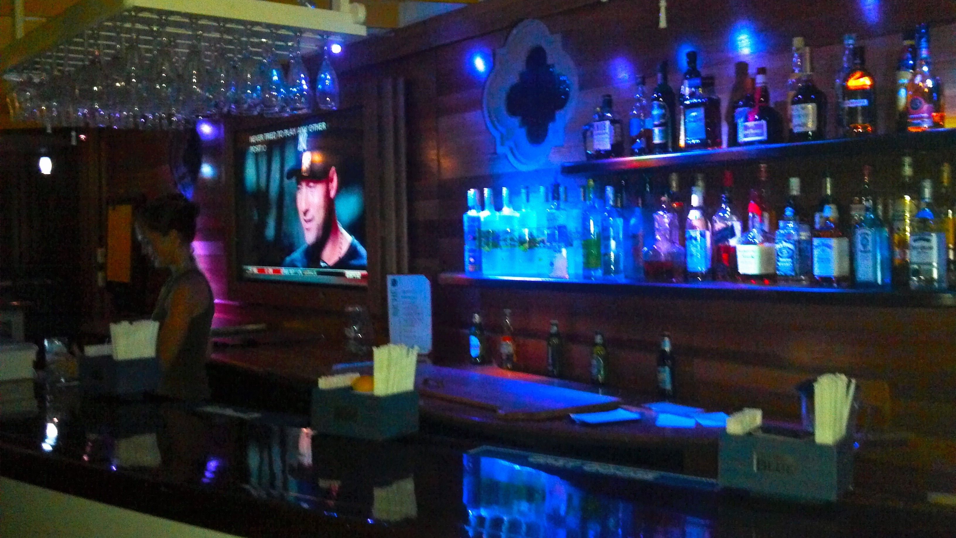 Zona de bar con una pantalla de televisión mostrando un programa, estanterías con varias botellas y una barra.