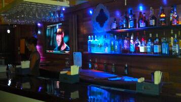 Zona de bar con una pantalla de televisión mostrando un programa, estanterías con varias botellas y una barra.