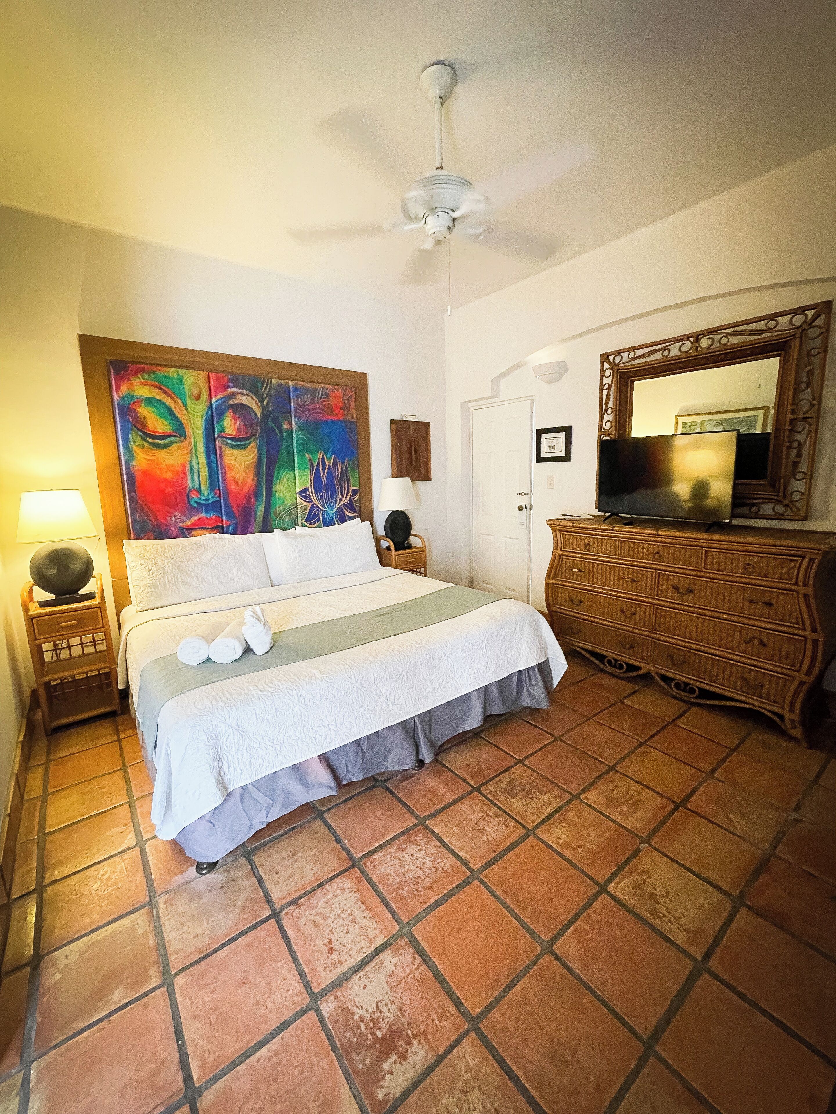A hotel room with a bed, a dresser, a television, a ceiling fan, a painting, and a lamp.