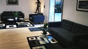 A modern living room with a sofa, a coffee table, and a statue. There is a window with a view of the city. - Hotel Ambassador Monaco (Monaco)