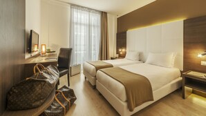 A hotel room with two beds, a desk with a chair, a TV, and a large bag on the floor. - Hotel Ambassador Monaco (Monaco)