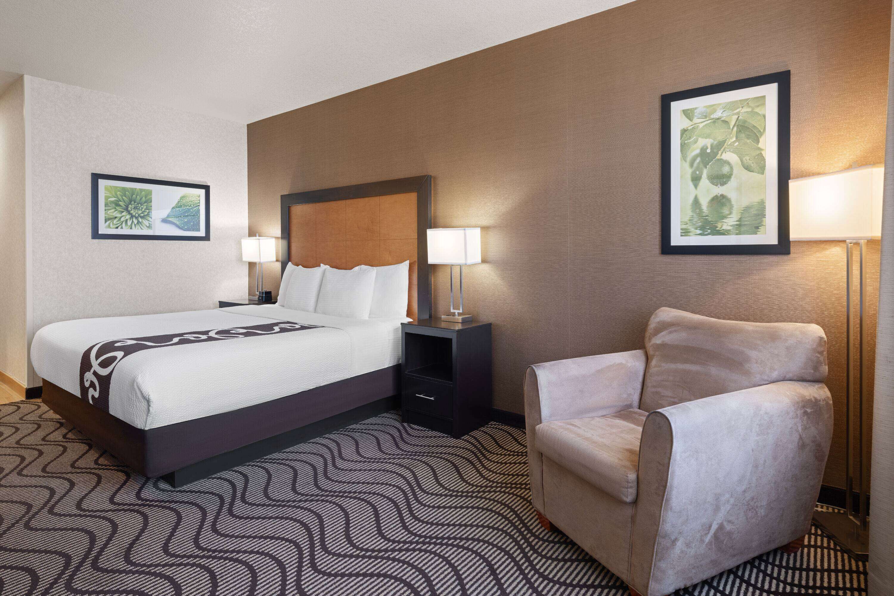 A hotel room with a bed, a nightstand, a chair, and two framed pictures on the walls.