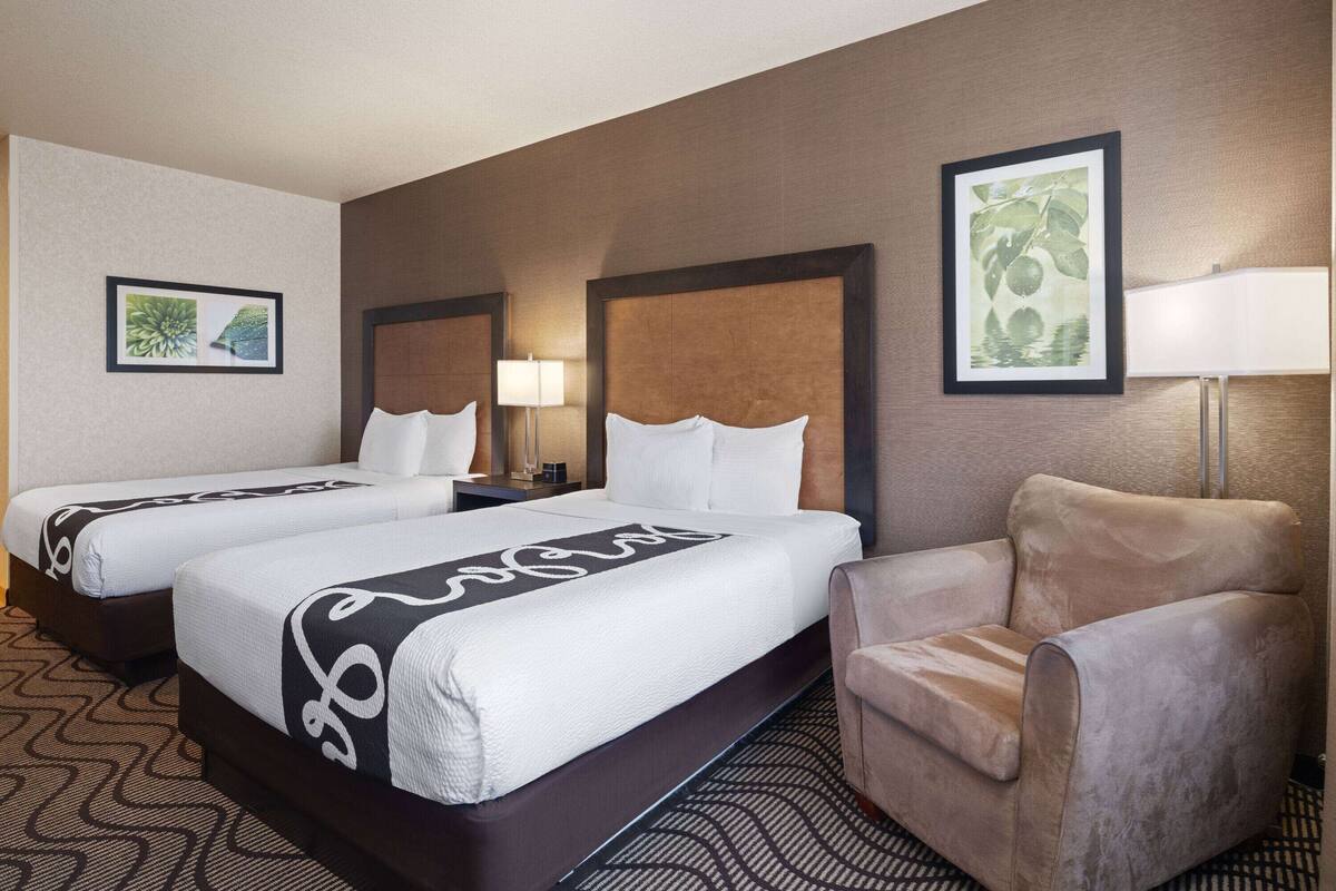 A hotel room with two beds, a chair, and a nightstand with a lamp.