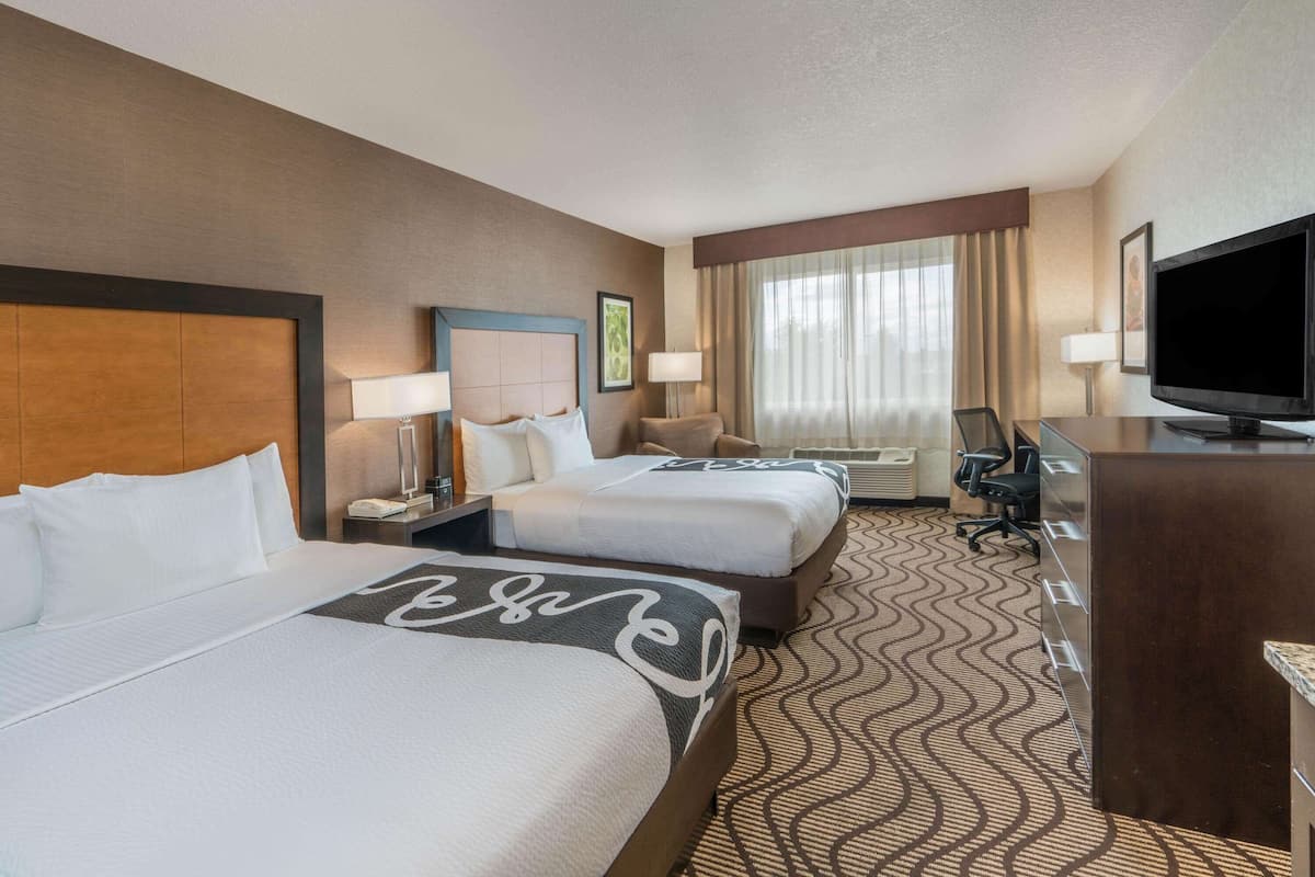 A hotel room with two beds, a desk, a television, and a large window with curtains.