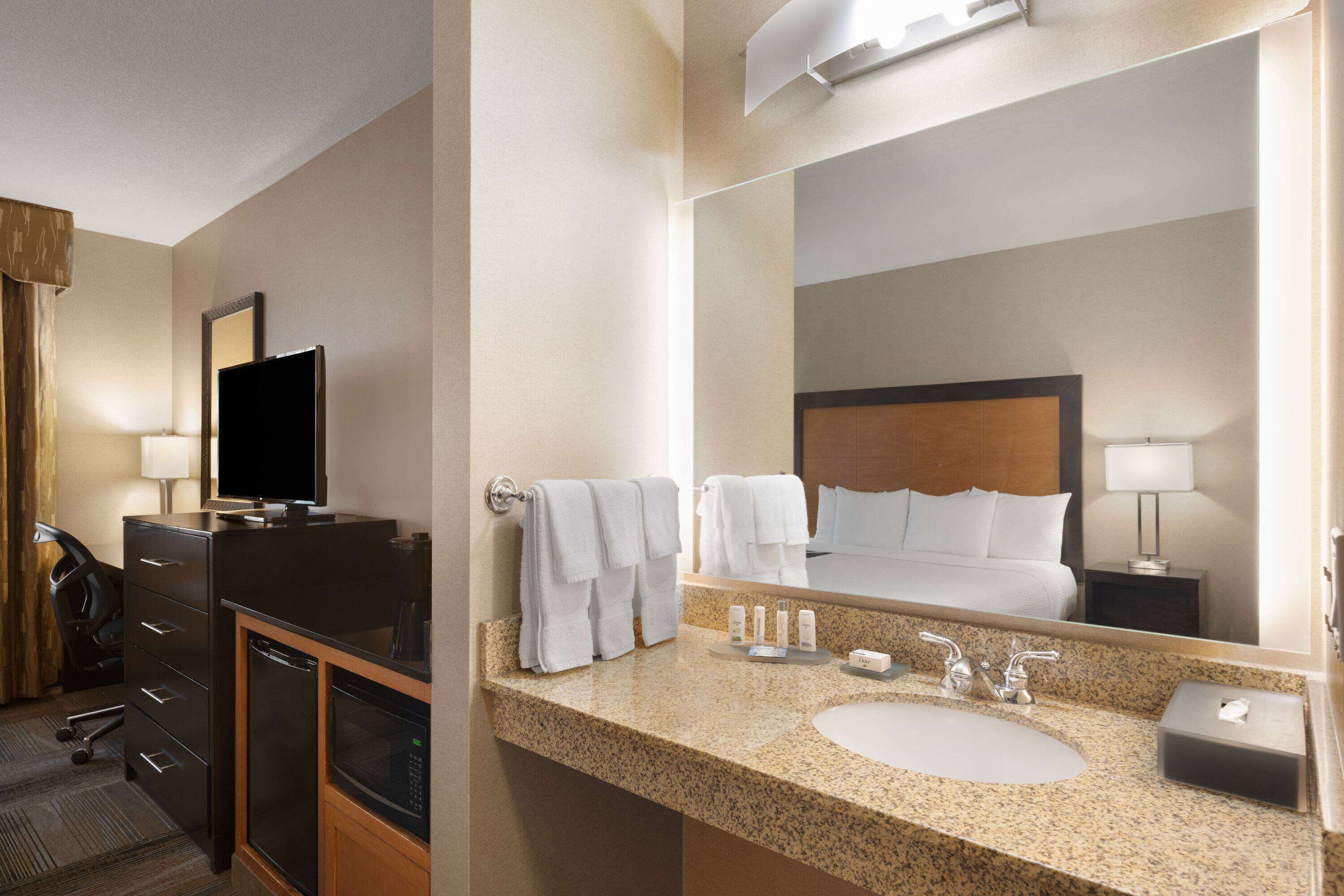 A hotel bathroom with a granite countertop sink, a mirror, a television, and a bed with white linens.
