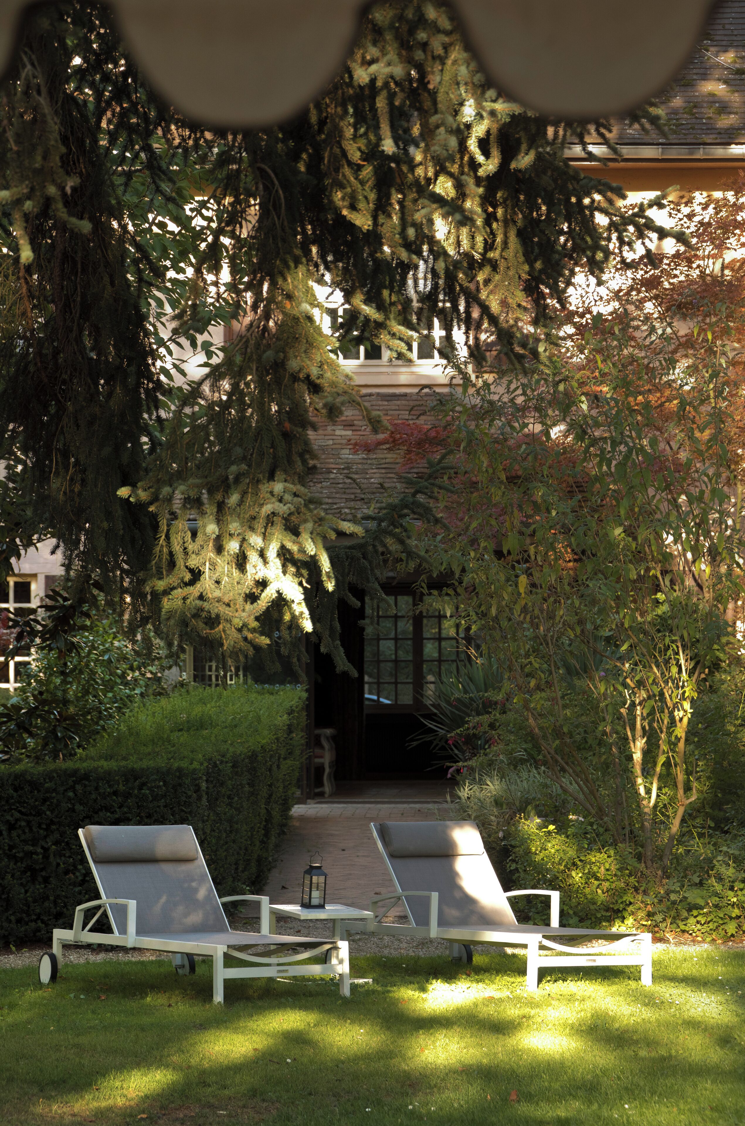 A garden with two lounge chairs and a table, surrounded by greenery and a building in the background.