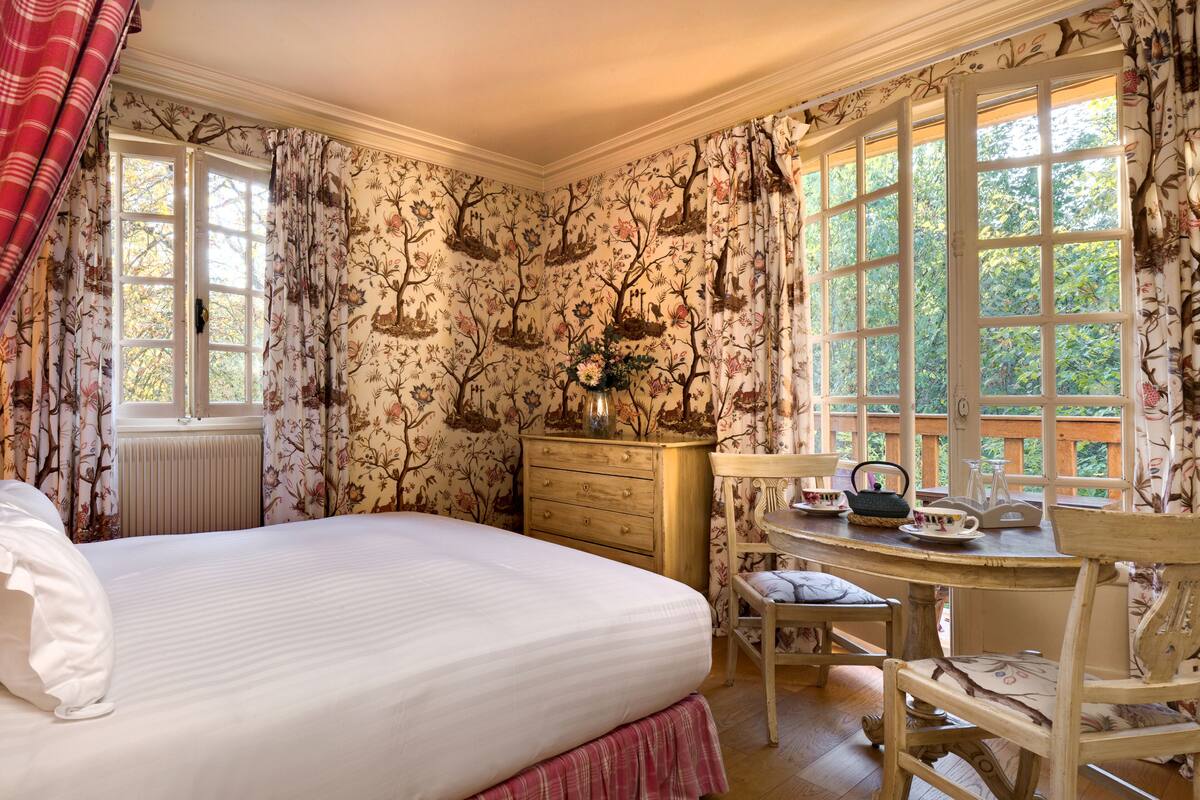 A bedroom with a large bed, a wooden dresser, a small table with chairs, and floral wallpaper.