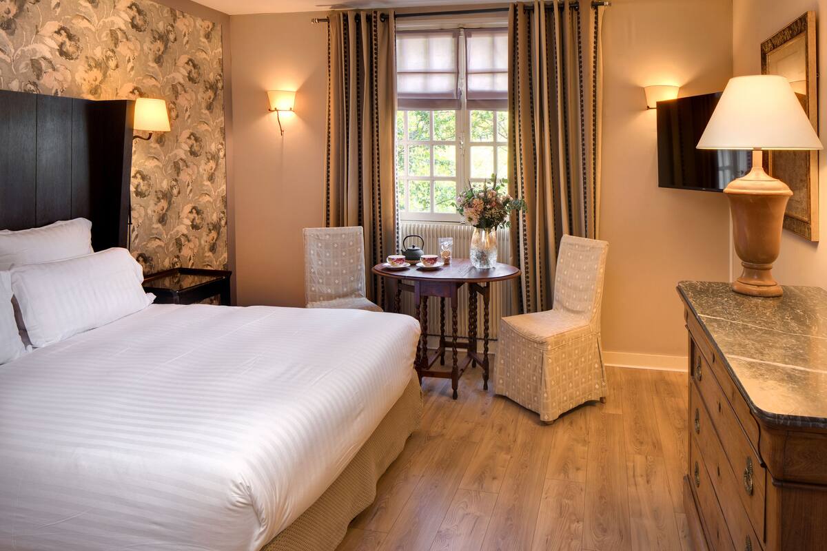 A hotel room with a large bed, a small table with a vase, two chairs, a dresser, and a window with curtains.
