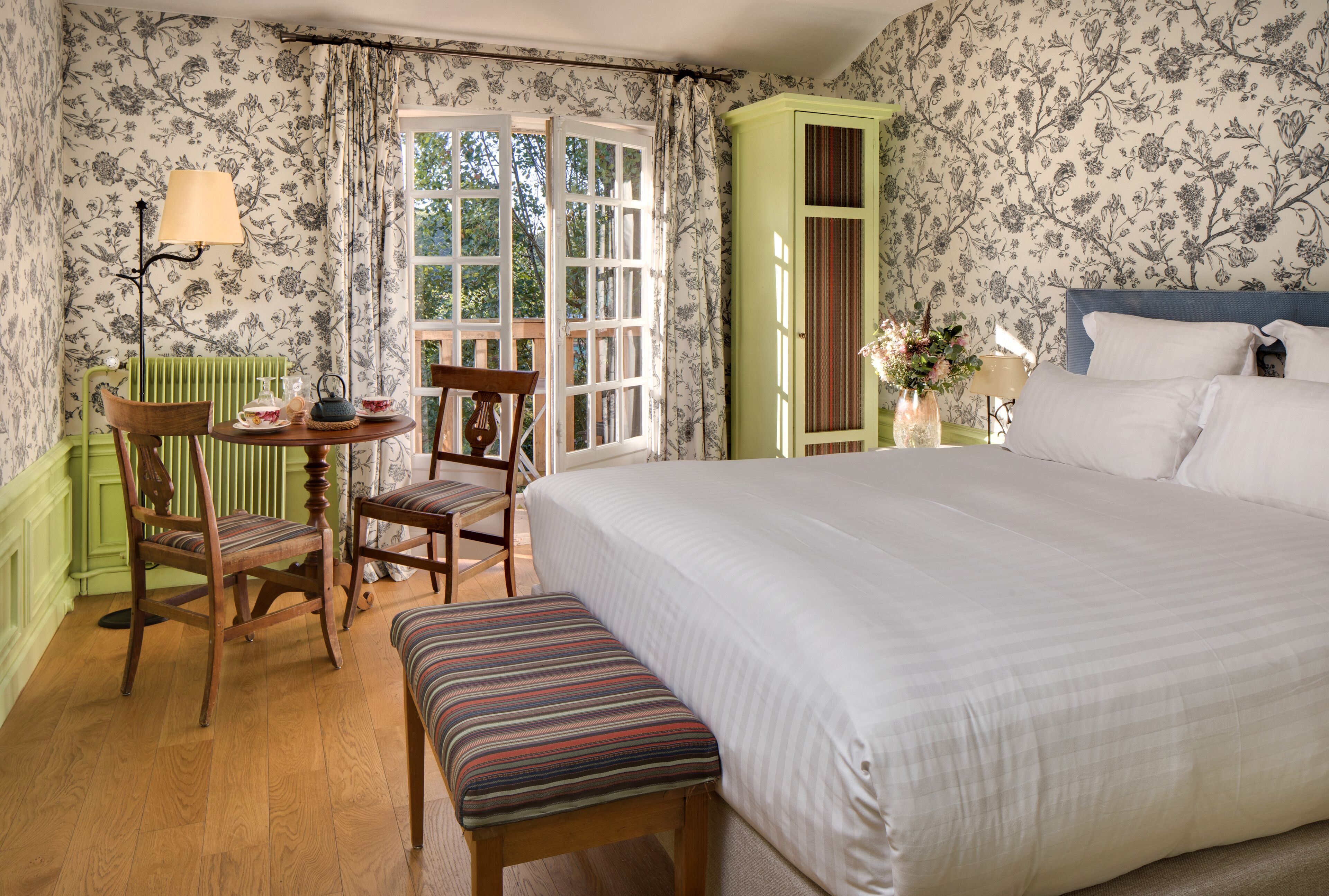 A bedroom with a large bed, a green cabinet, a small table with chairs, and a window with curtains.