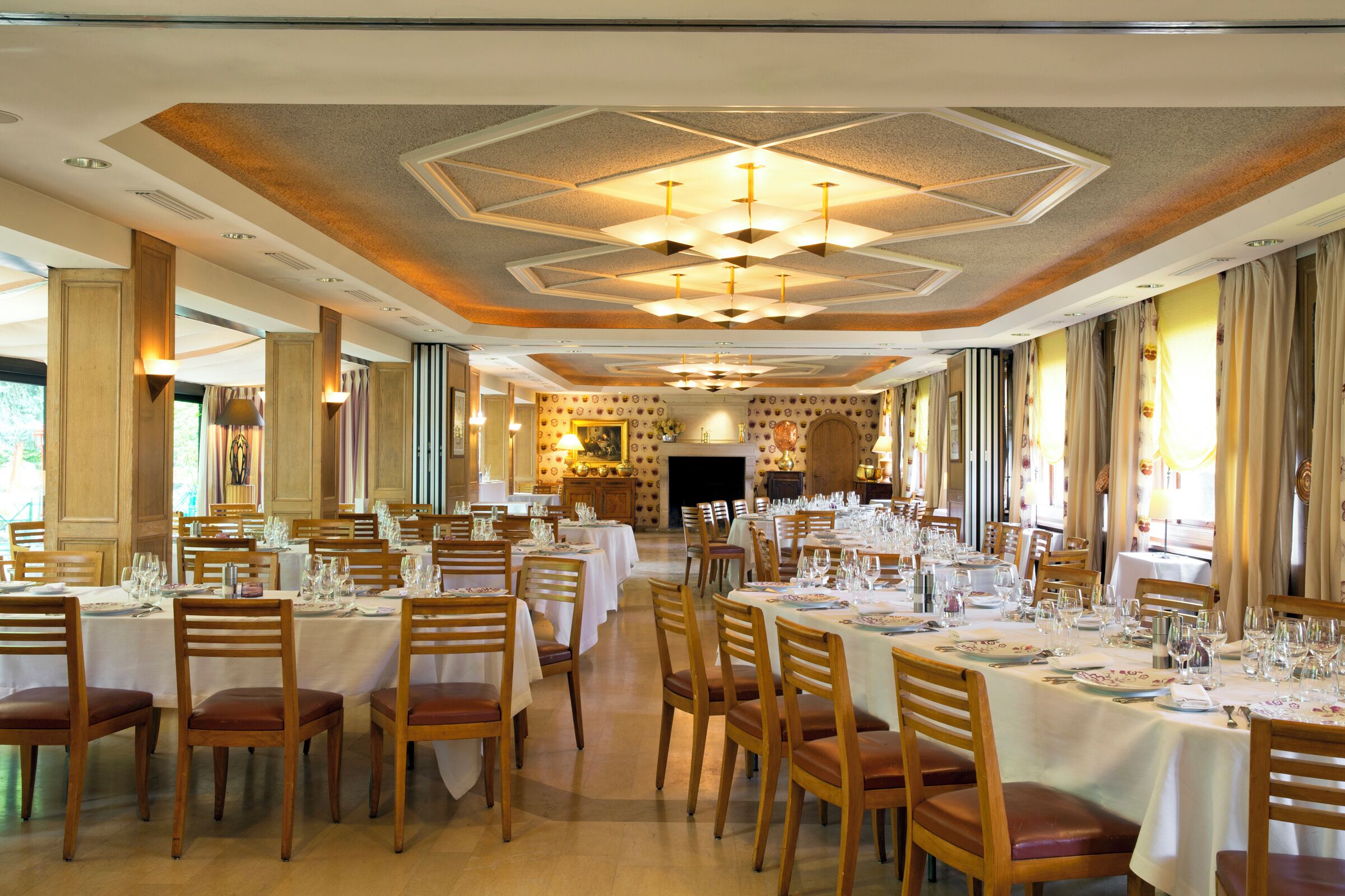 A large dining hall with multiple tables set for a meal, wooden chairs, and a decorative ceiling.
