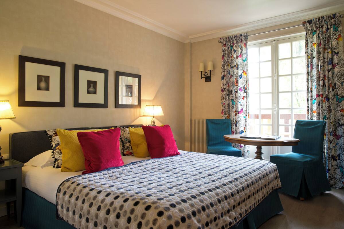 A hotel room with a bed, two side tables, a chair, a small table, and a window with curtains.