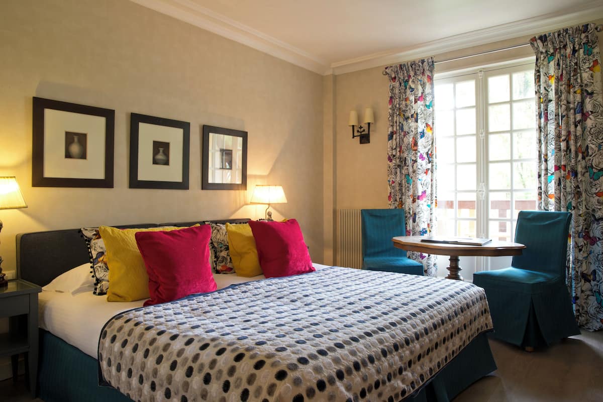 A hotel room with a bed, two side tables, a chair, a small table, and a window with curtains.