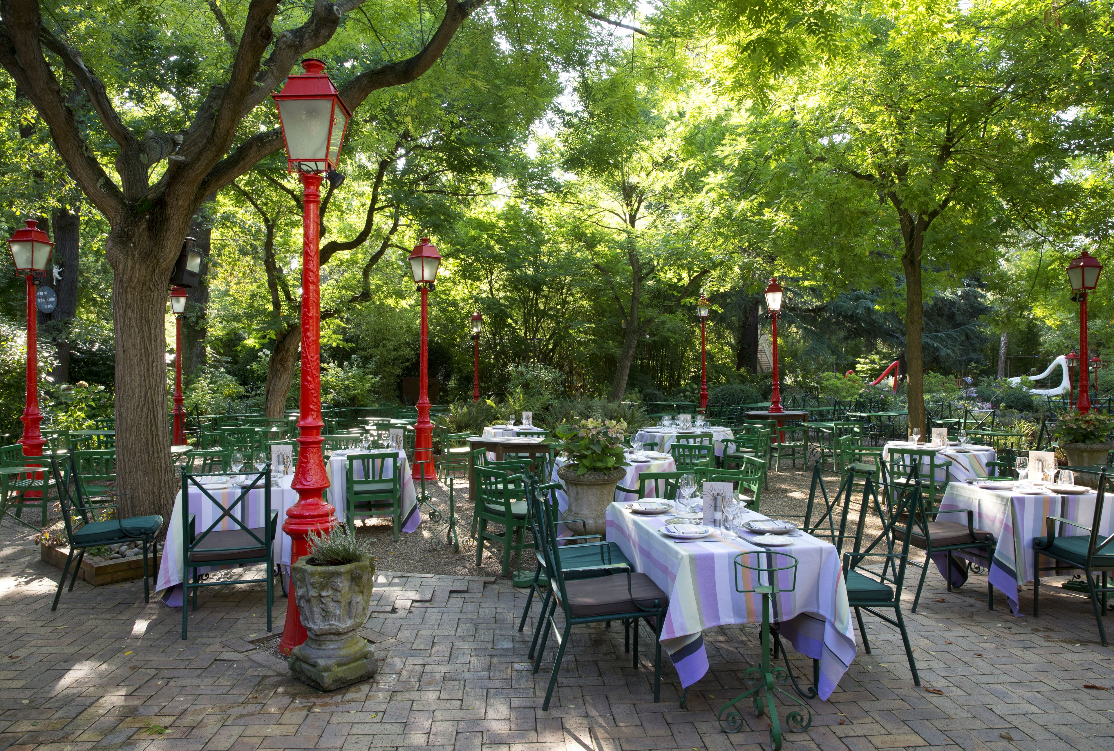 An outdoor dining area with tables and chairs, surrounded by trees and red street lamps.