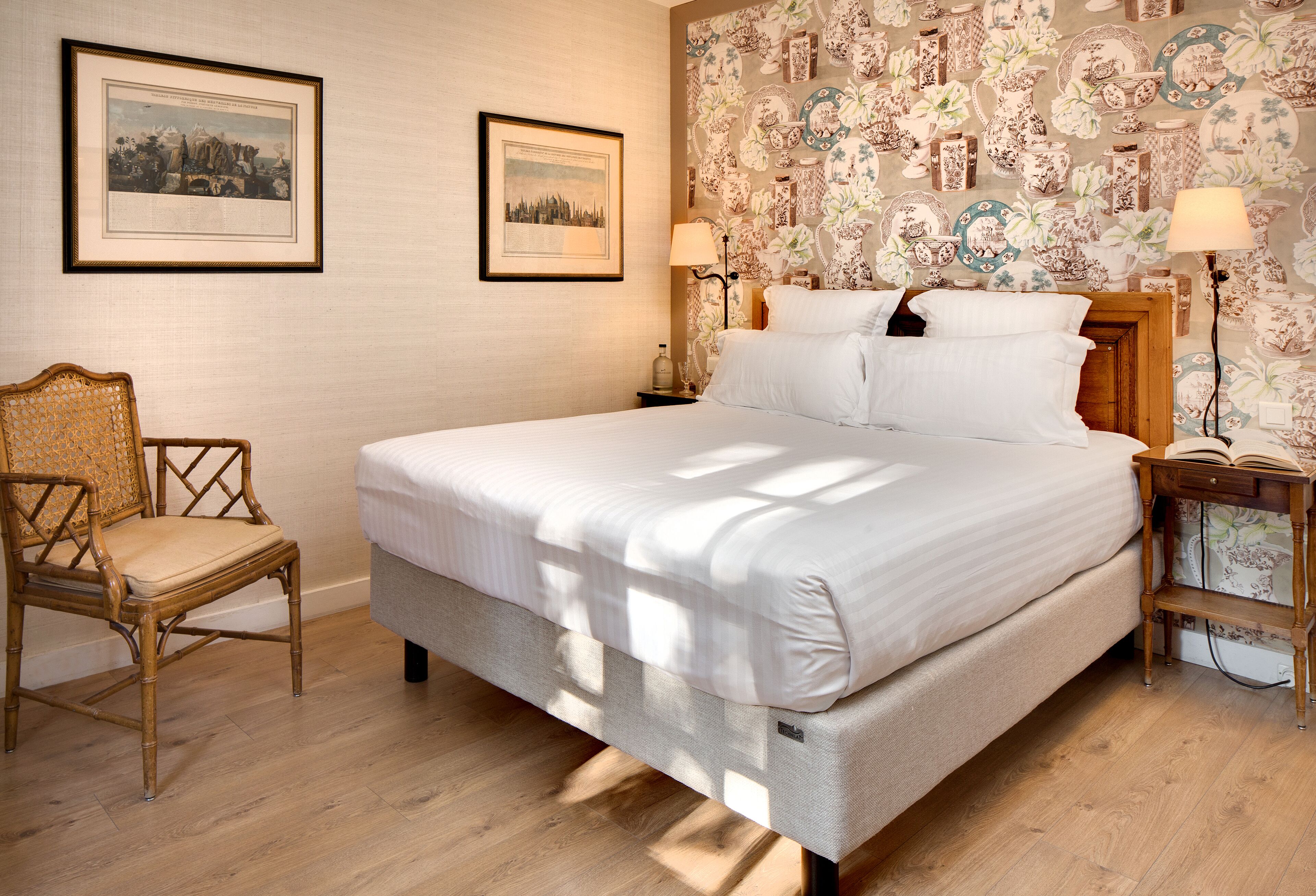 Un dormitorio ordenado con una cama grande, dos mesitas de noche, una silla y una pared decorada con cuadros.