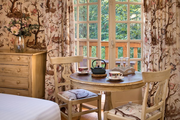 A room with a wooden dresser, a round table with chairs, a vase with flowers, and a window with a view of greenery.