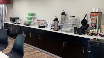 A modern kitchen with dark cabinets, a white countertop, and a coffee machine. There are two black chairs and a table.