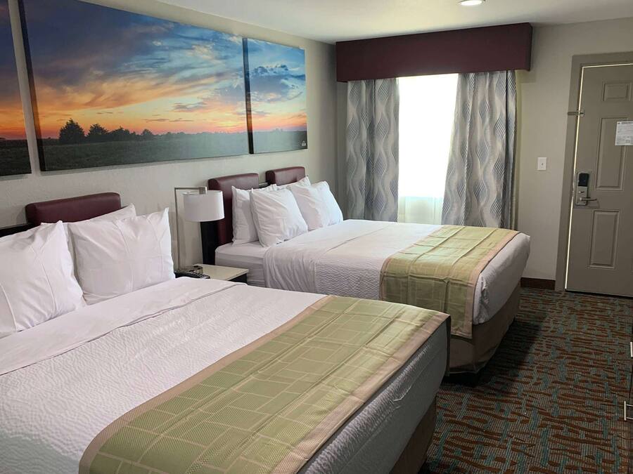 Een hotelkamer met twee bedden, een groot schilderij van een zonsondergang, een raam met gordijnen en een deur met een bordje.