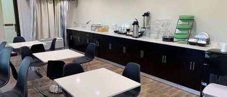 A modern hotel kitchen with white tables, black chairs, and a counter with various appliances and supplies.
