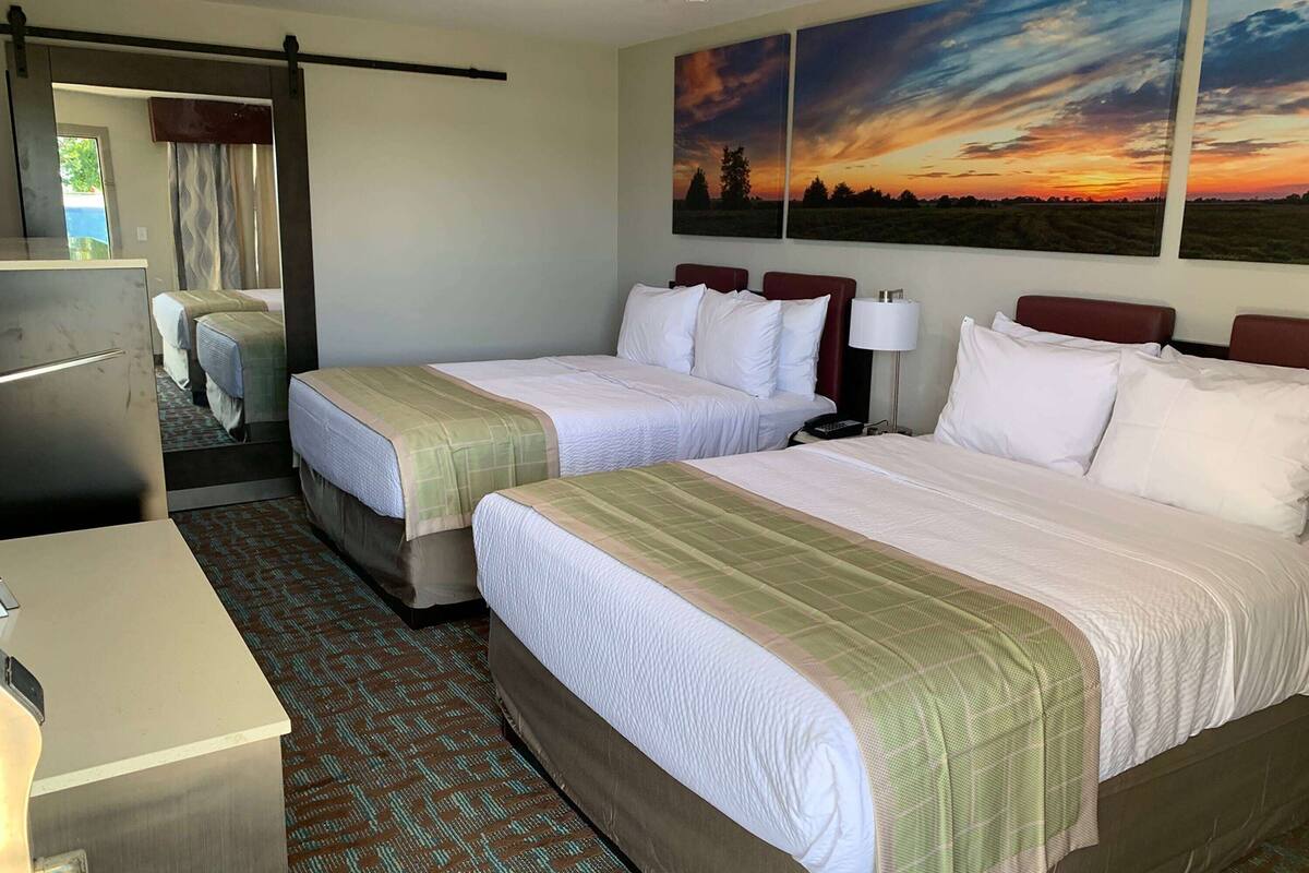 A hotel room with two beds, a desk, a mirror, and a large window with a sunset view.