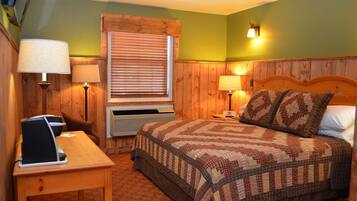 A bedroom with a wooden headboard, a quilted bedspread, two bedside lamps, a window with blinds, and a desk with a laptop.