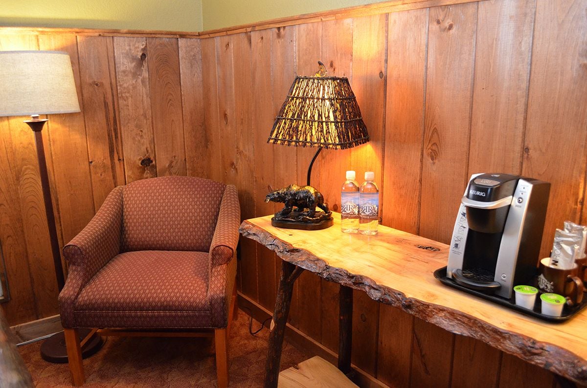 A cozy corner with a patterned armchair, a rustic wooden table with a coffee maker, a lamp, and two bottles of water.