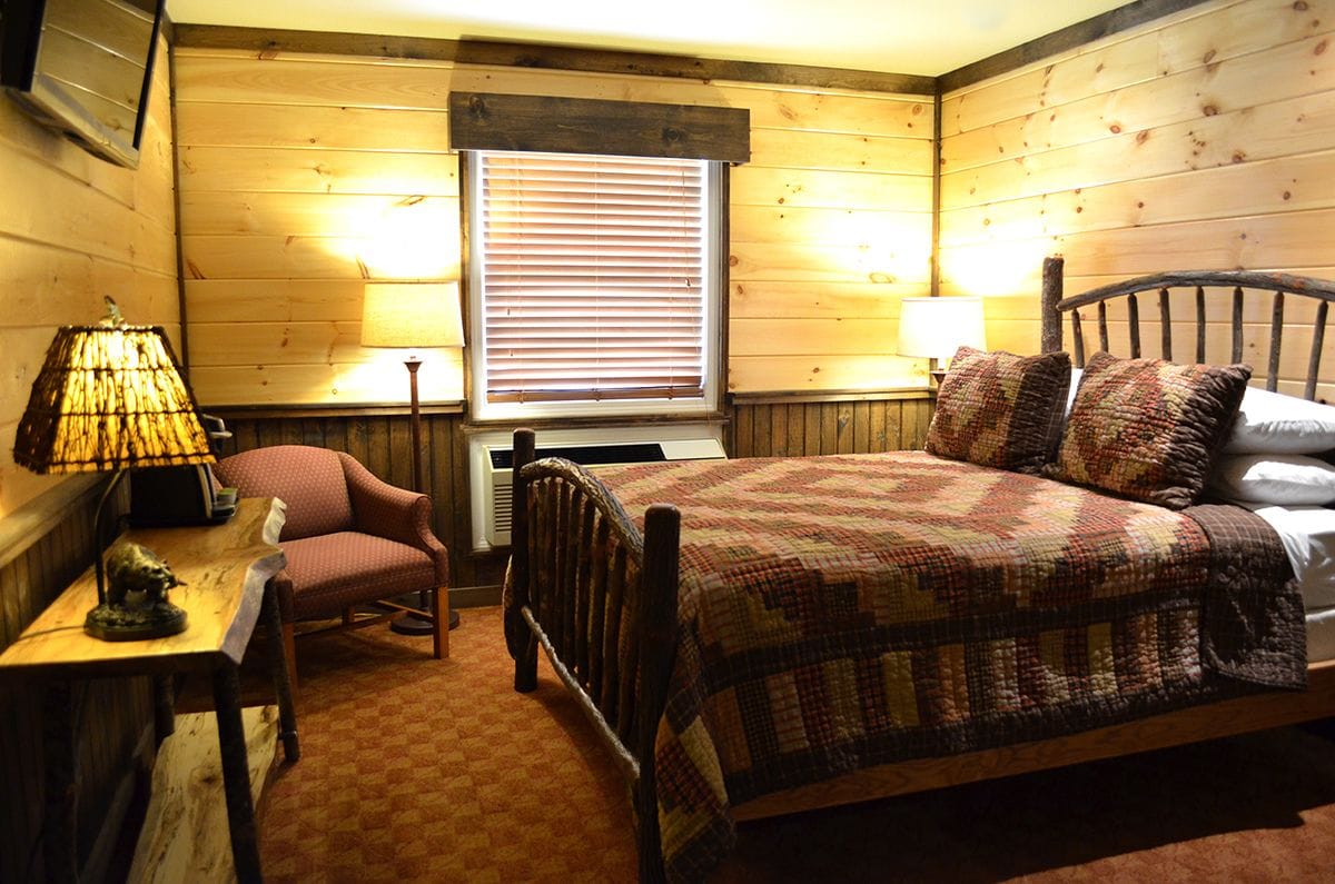 A cozy bedroom with a wooden bed, a chair, a table with a lamp, and a window with blinds.