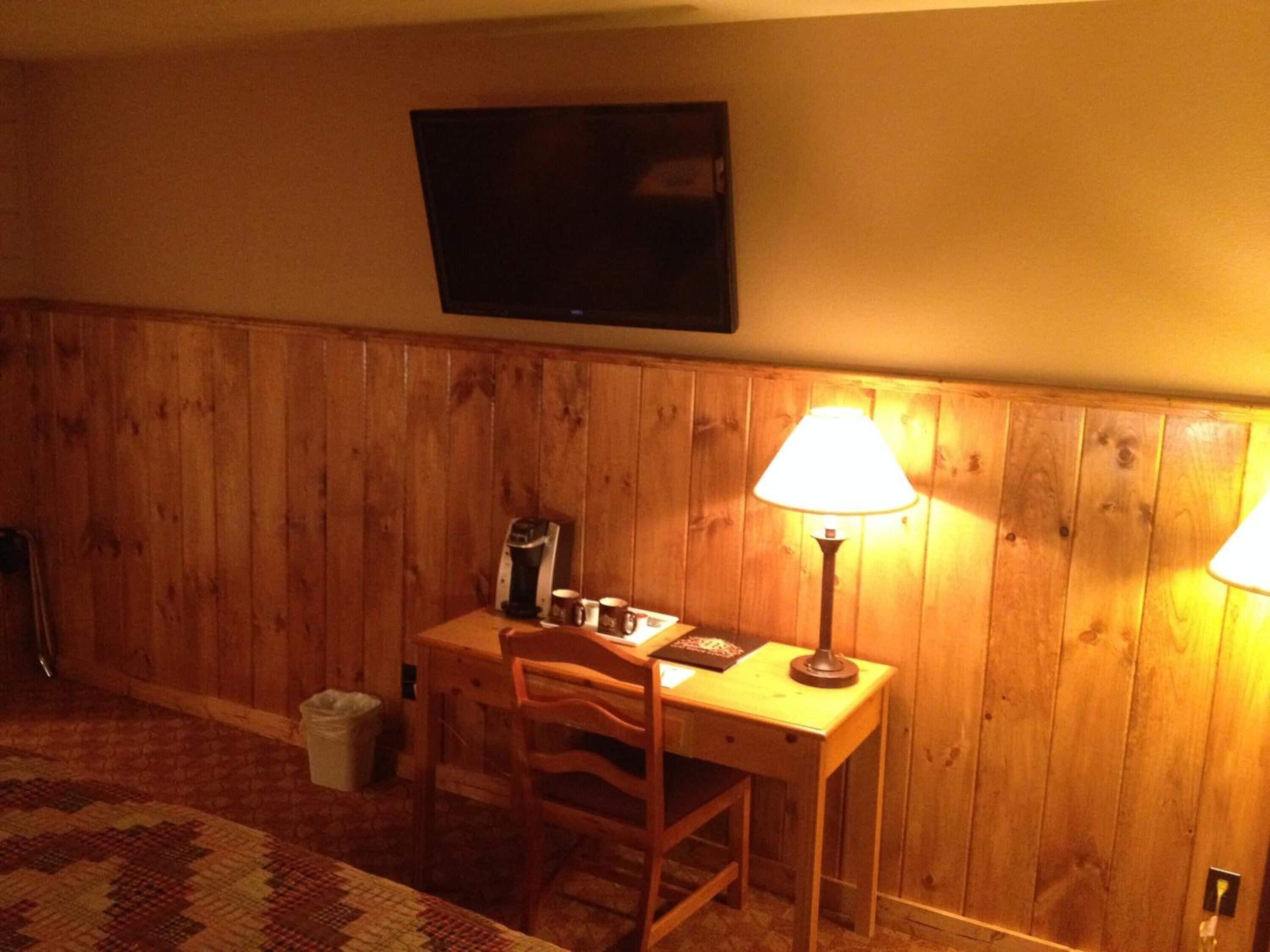A room with wooden panel walls, a television mounted on the wall, a desk with a lamp, and a chair.