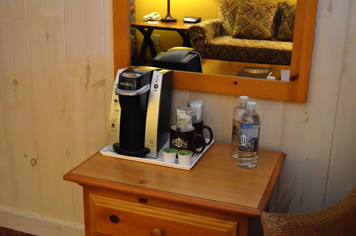 Une machine à café, une tasse et une bouteille sont posés sur une table en bois dans une pièce où un miroir reflète un canapé et une lampe.