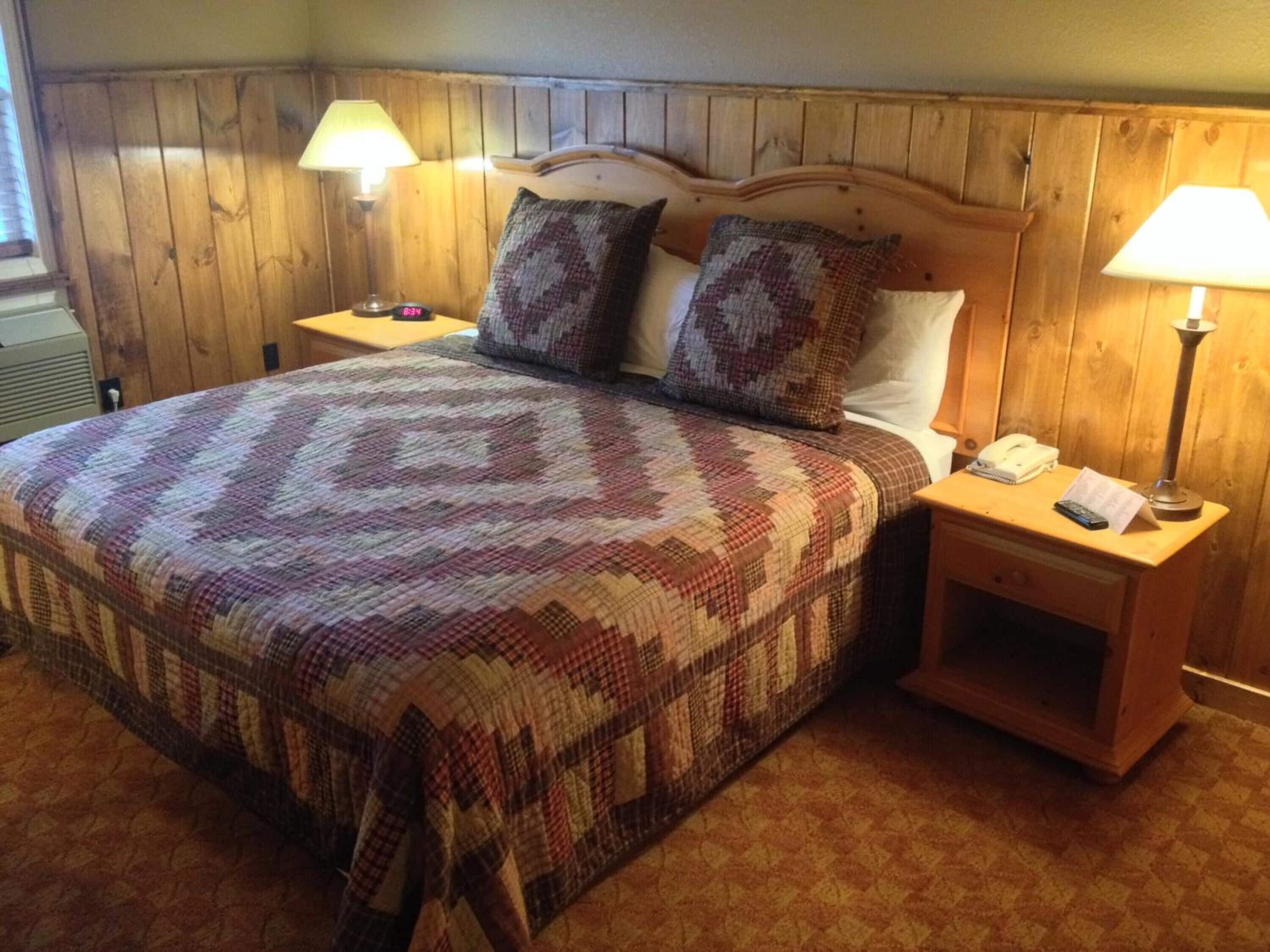 A hotel room with a wooden headboard, a large bed with a patterned blanket, two bedside lamps, and a nightstand with a phone and a lamp.