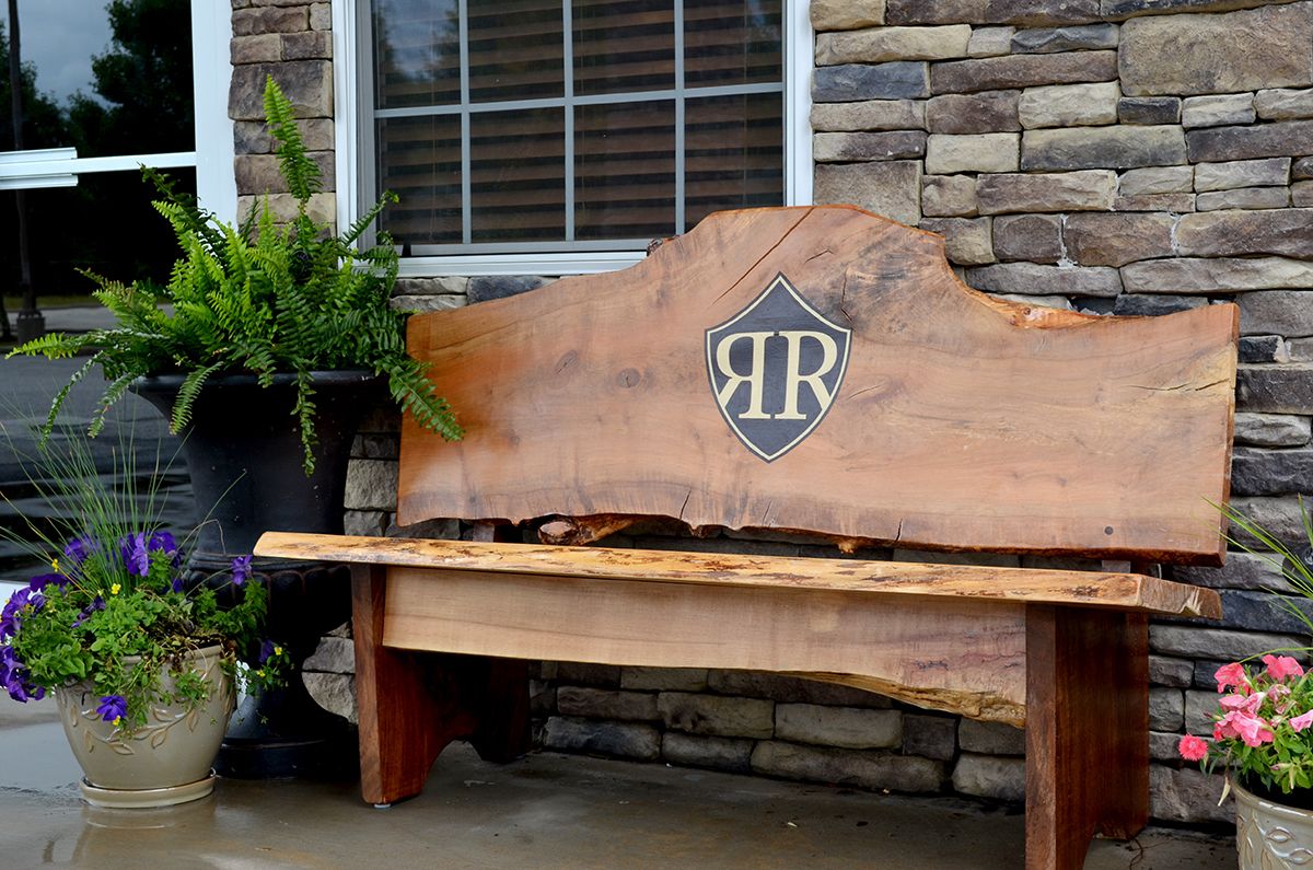 A rustic wooden bench with a shield-shaped plaque bearing the initials "AR" against a stone wall backdrop.
