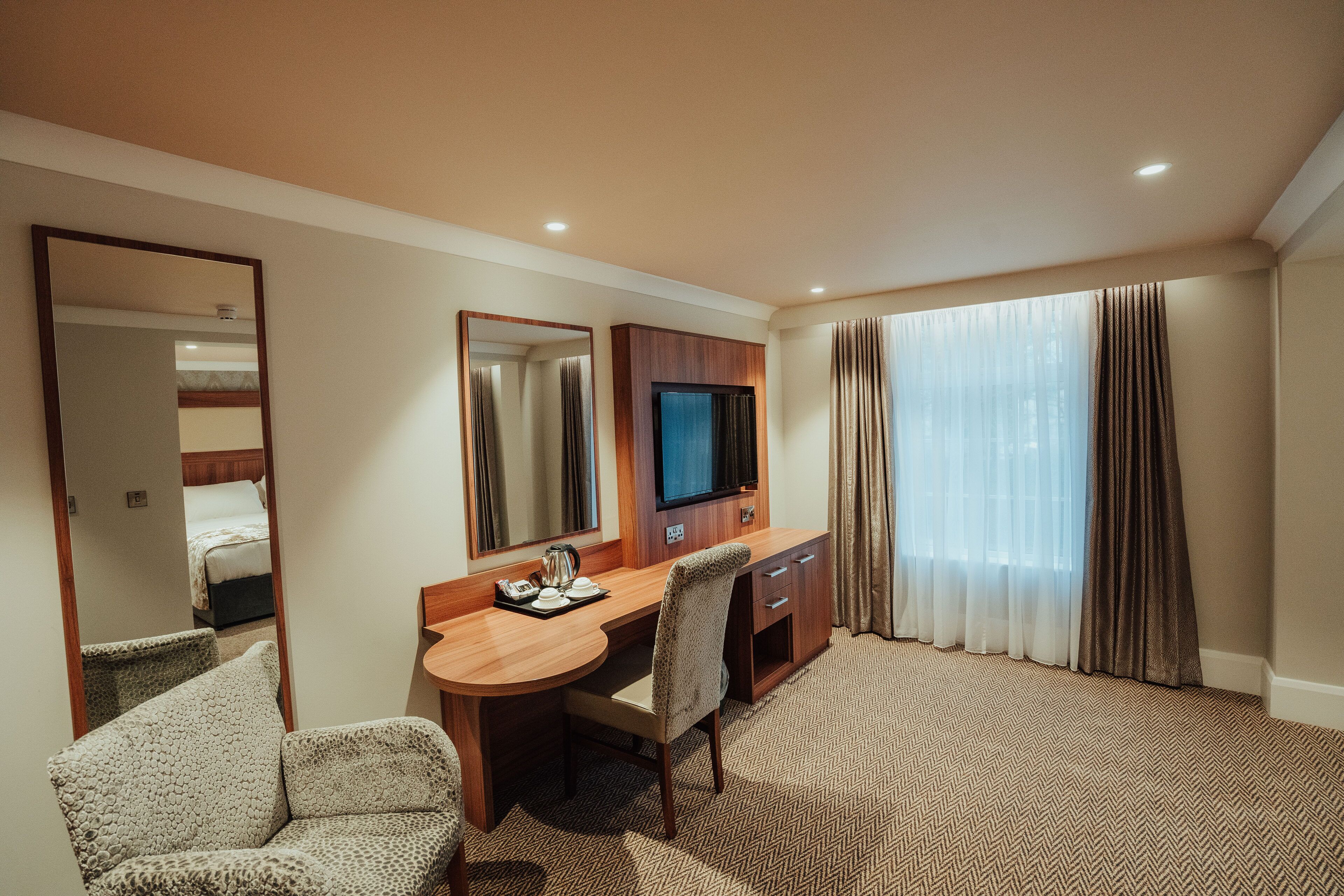 Habitación de hotel con un escritorio de madera, una silla, un televisor y un ventanal con cortinas.