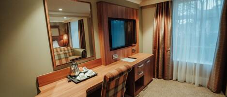 A hotel room with a wooden desk, a television, a mirror, a chair, and a window with curtains.