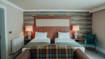 A hotel room with two beds, a wooden headboard, two bedside lamps, a plaid throw blanket, and a green armchair.