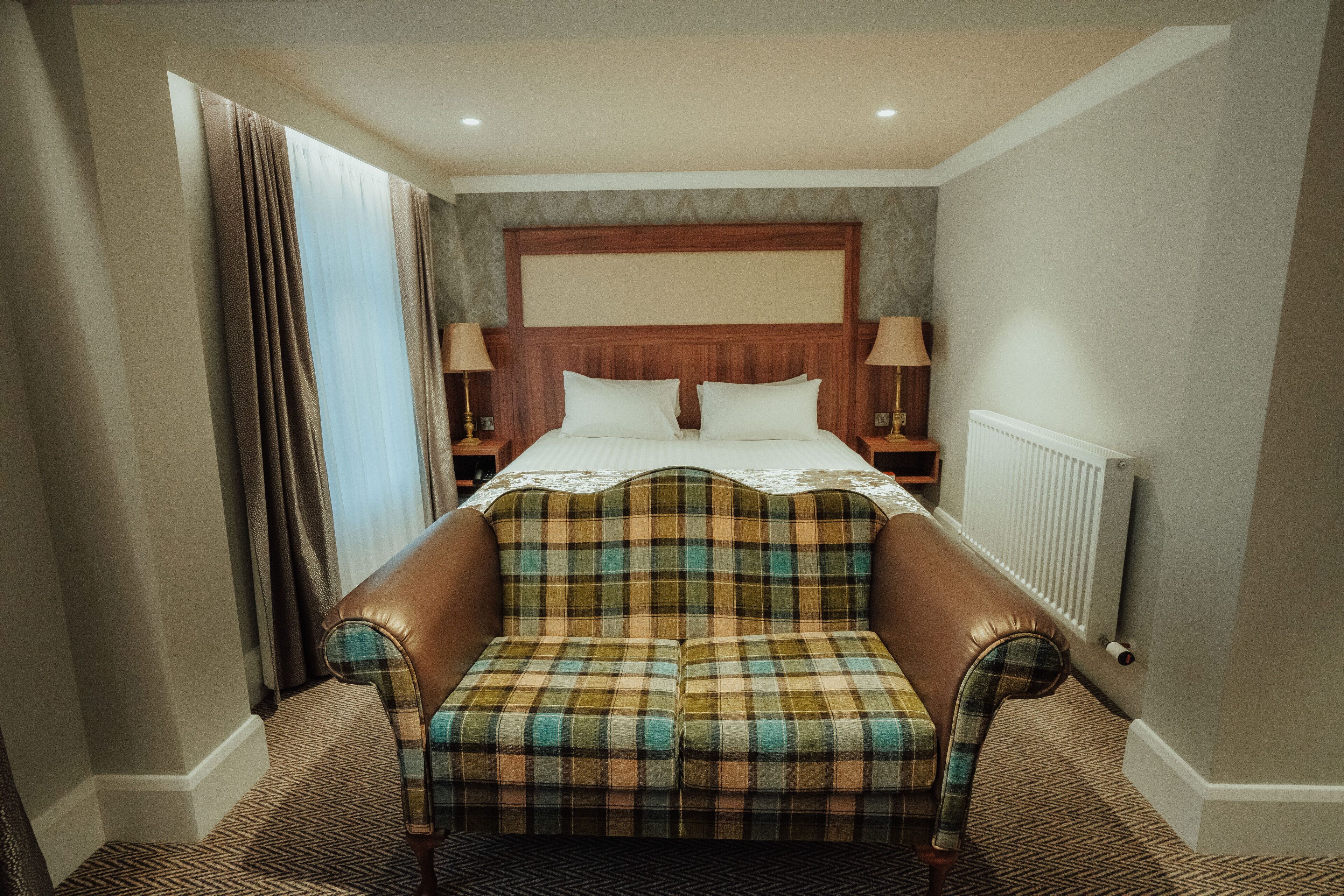 Une chambre d’hôtel avec un grand lit, deux tables de chevet, une tête de lit, une fenêtre avec des rideaux et un canapé.