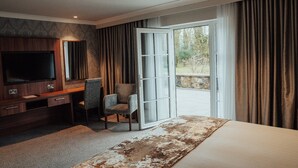 A hotel room with a large bed, a desk with a TV, a chair, and a view of a garden through an open door. - Dunadry Hotel and Gardens (Antrim)